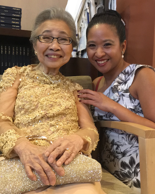 An older southeast asian woman wearing a gold dress is smiling while sitting in a chair. Next to her adult granddaugther wearing a floral dress and she is also smiling. 
