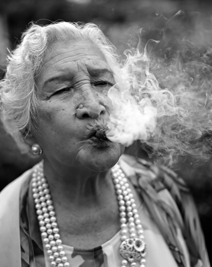 A black and white photo of an southeast asian woman smoking a cigar. Her hair and dress are styled in an 1930s fashion. 